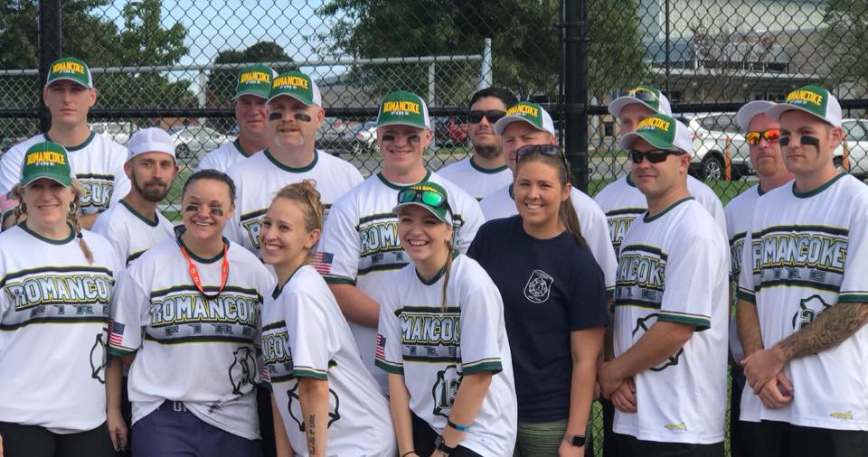 A coed softball team wearing matching "ROMANOCKO" white jerseys and green hats poses together outside on a sunny day in front of a chain-link fence.