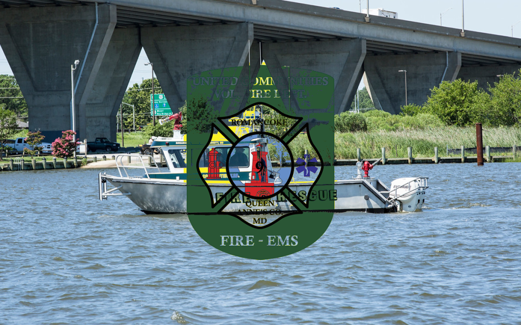 A small rescue boat with two people is on the water near a large bridge. There is a green fire and rescue badge from Kent Island Volunteer Fire Department overlaid on the image.