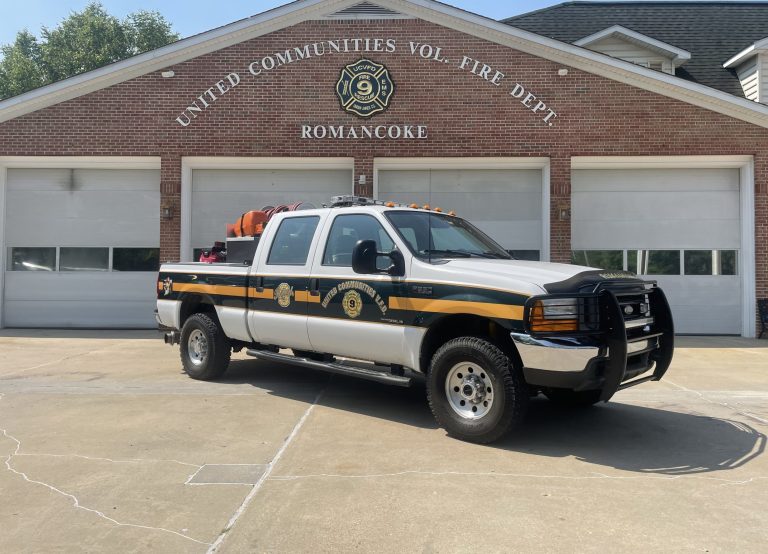 A white and green fire department pickup truck is parked in front of the United Communities Volunteer Fire Department building in Romancoke, with three garage doors in the background.