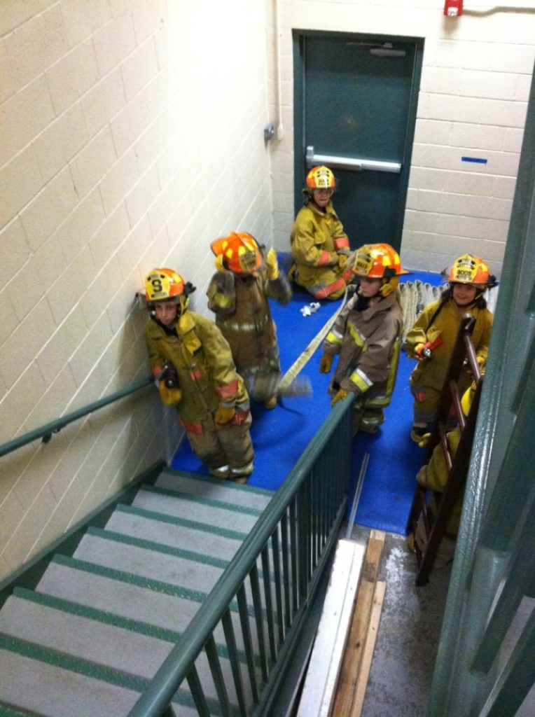Five children dressed in firefighter gear stand in a stairwell next to a door, holding hoses and equipment, appearing to participate in a fire safety drill or training exercise.