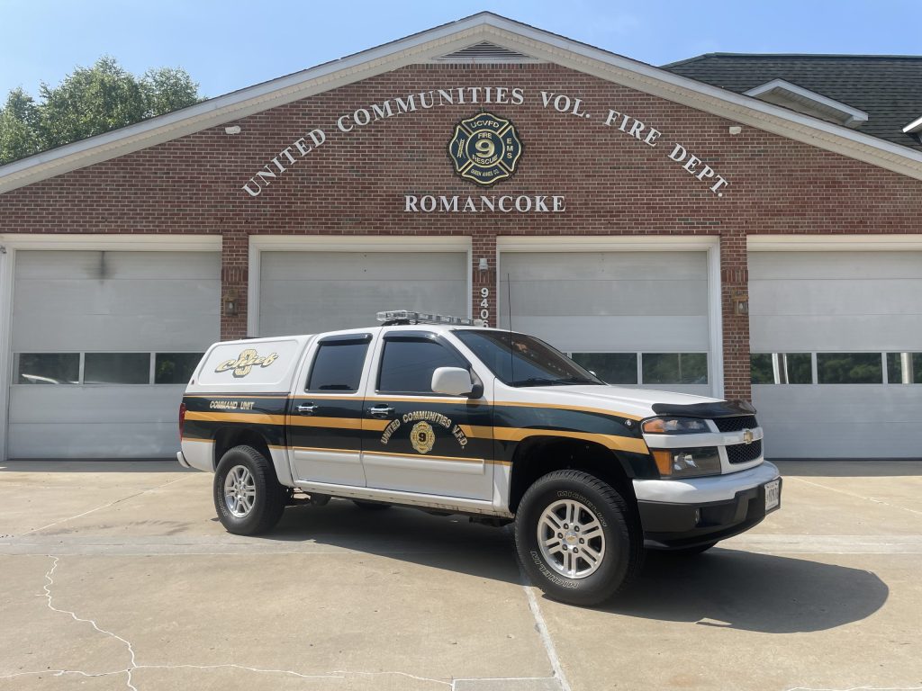 A black and white emergency response truck labeled "Command Unit" is parked in front of a brick fire station with the sign "United Communities Vol. Fire Dept. Romancoke" above the garage doors.