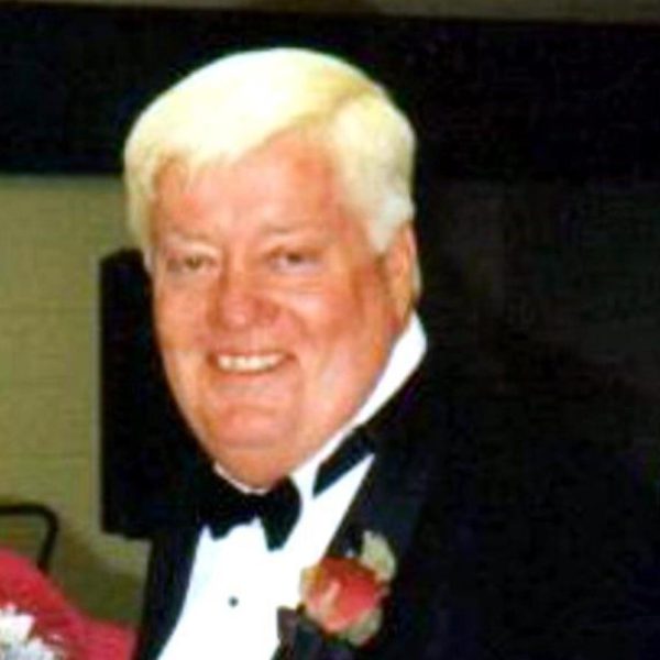 A smiling man with short, white hair wears a black tuxedo, white shirt, and black bow tie. A red rose boutonniere is pinned to his lapel. The background is out of focus and appears to be indoors.