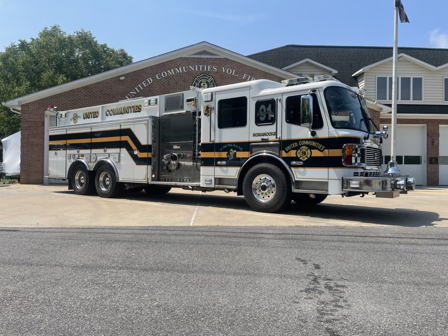 A large white and black fire and rescue truck is parked in front of a brick fire station with the words "United Communities Vol. Fire" on the building and the truck. The scene is sunny with trees in the background.