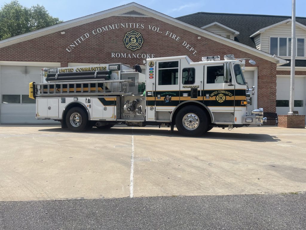A black, white, and yellow fire truck is parked in front of the United Communities Volunteer Fire Department building in Romancoke on a sunny day. The firehouse has brick walls and a peaked roof.