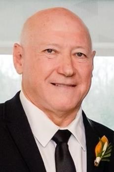 A bald man in a black suit, white shirt, and black tie, smiling at the camera. He has a boutonniere with orange petals on his lapel and is standing indoors with a blurred background.