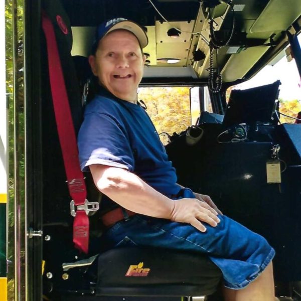 A smiling man in a blue shirt, denim shorts, and a baseball cap sits in the driver's seat of a large vehicle, possibly a fire truck, with control panels and equipment visible around him.