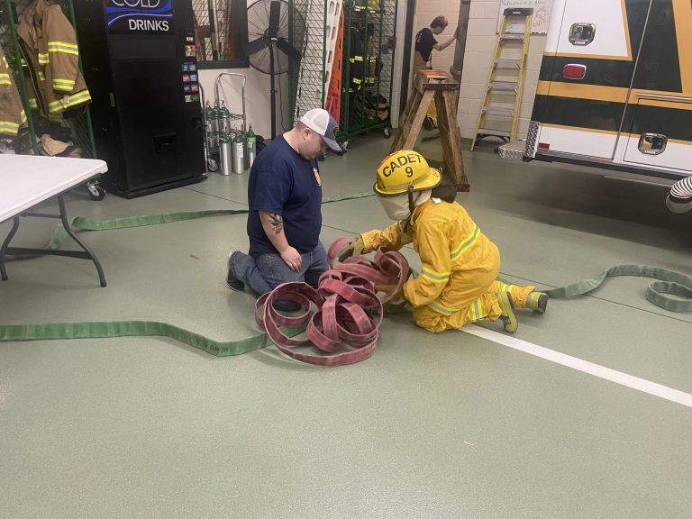 A firefighter and a cadet in yellow gear kneel on the floor, practicing with coiled fire hoses inside a fire station next to a table and emergency vehicle.