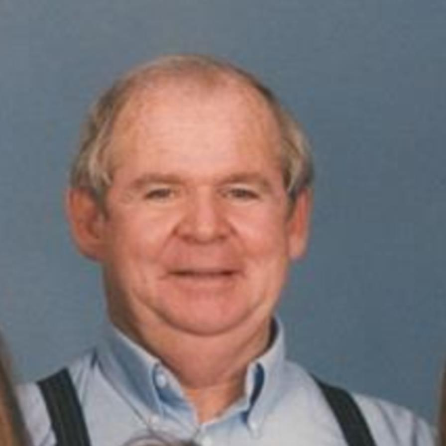 An older man with short gray hair wearing a light blue button-up shirt and dark suspenders, smiling in front of a plain blue background.