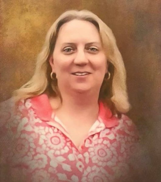 A woman with long blonde hair is smiling, wearing hoop earrings and a pink floral patterned top, posed in front of a softly blurred brown background.