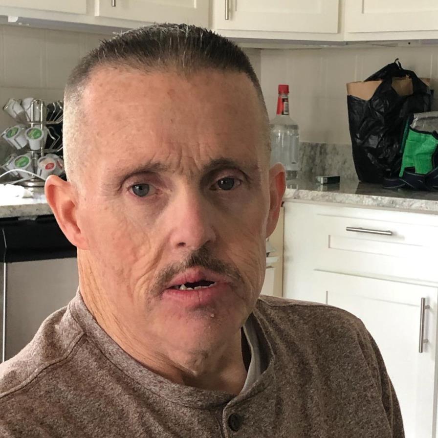 A middle-aged man with short hair and a mustache sits in a kitchen, wearing a brown buttoned shirt. There are cabinets, a countertop, and various kitchen items visible in the background.