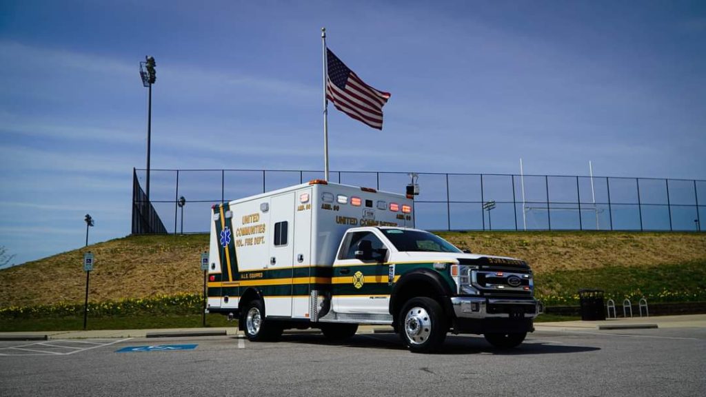 A white and green ambulance is parked in an empty lot under a clear blue sky. An American flag is flying on a pole behind the vehicle, near a grassy hill and a sports field with goalposts.