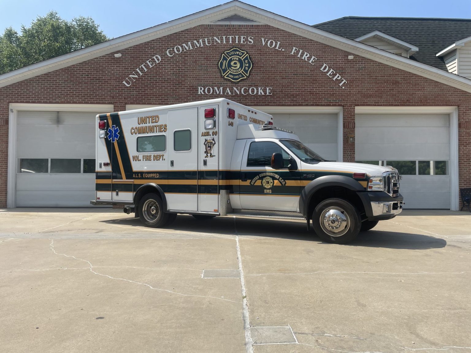 A white and green ambulance is parked in front of a brick fire station with "United Communities Vol. Fire Dept. Romancoke" written above three garage doors.