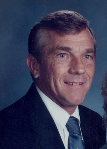 A middle-aged man with short gray hair and blue eyes is wearing a dark suit, white shirt, and patterned blue tie, smiling slightly against a plain blue background.