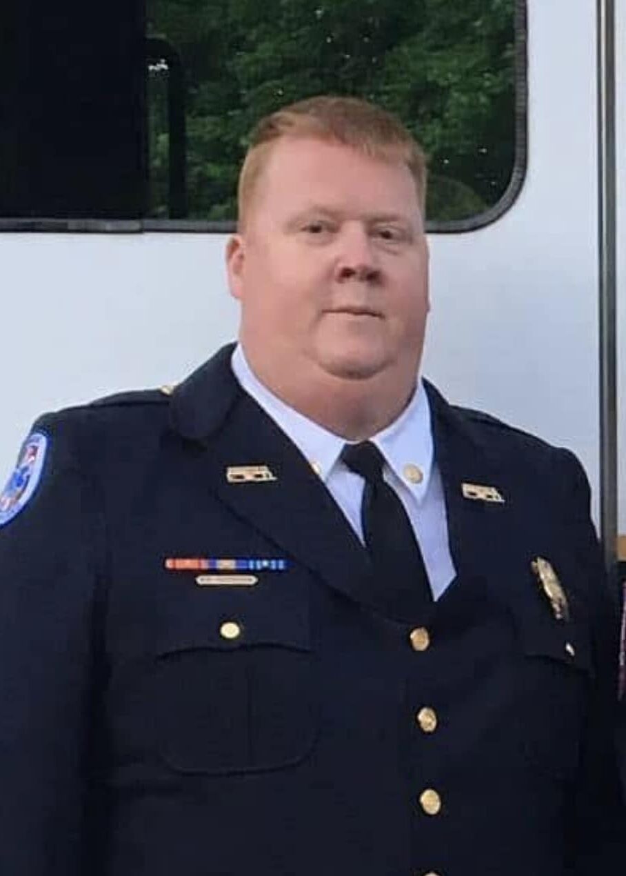 A man with short red hair wearing a dark blue formal uniform, badge, and tie stands in front of a white vehicle, with greenery reflected in a window behind him.