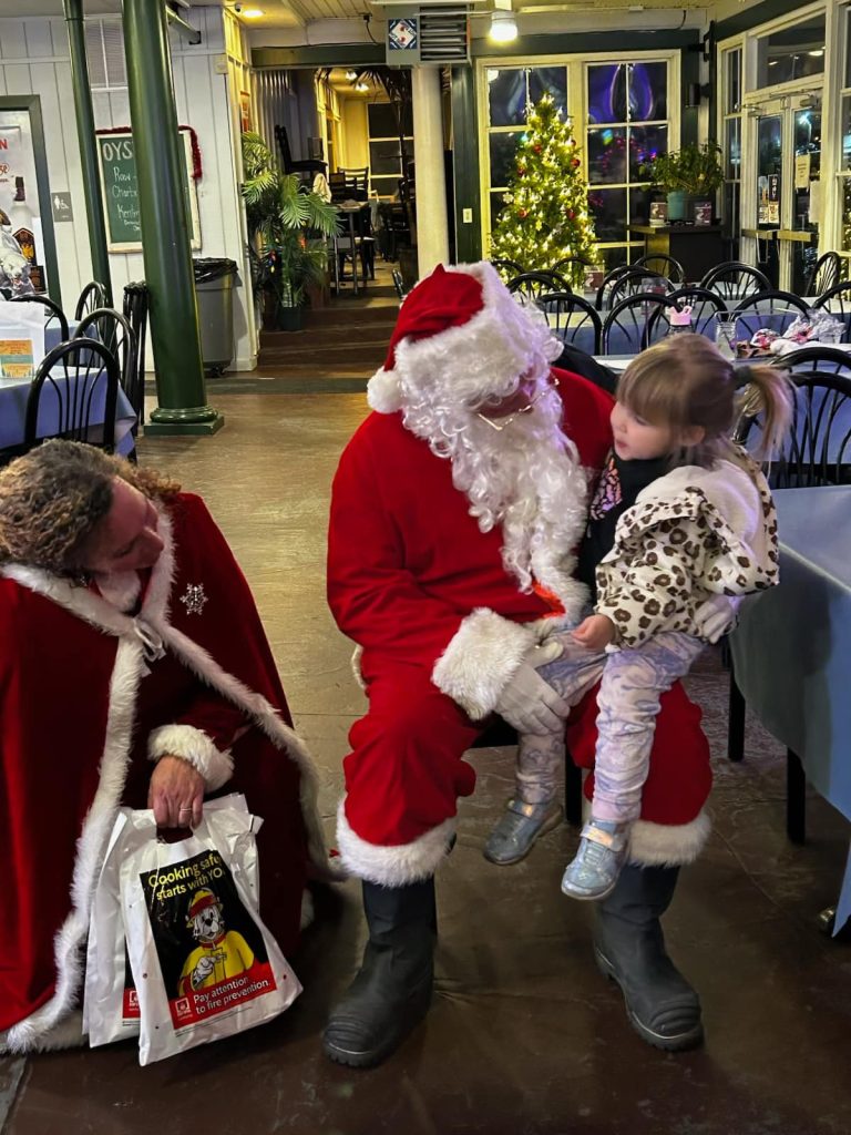 A child sits on Santa's lap while another person in a Santa cape kneels nearby holding a gift bag. They are indoors, with holiday decorations and a Christmas tree in the background.