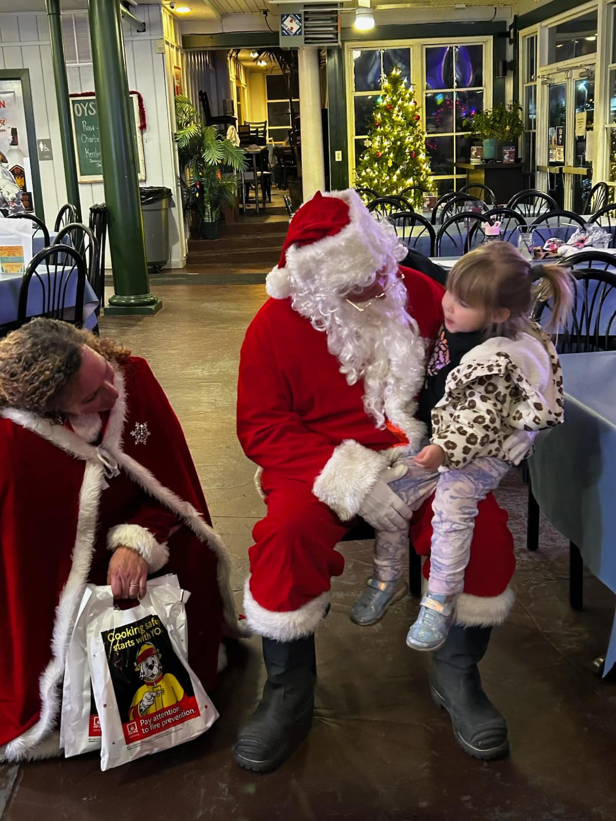 A child sits on Santa's lap while another person in a Santa cape kneels nearby holding a gift bag. They are indoors, with holiday decorations and a Christmas tree in the background.