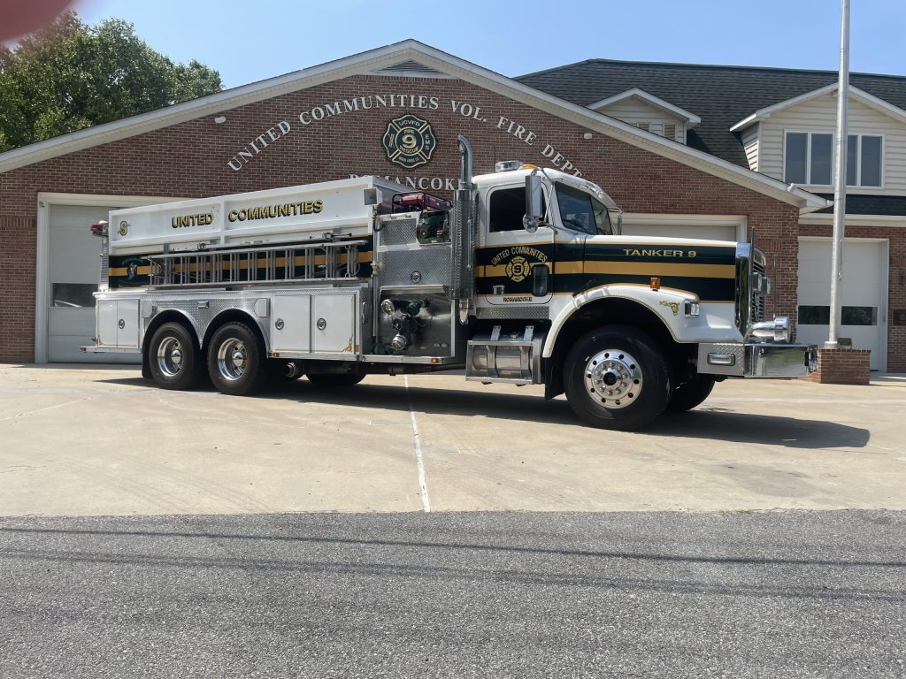 A large white and green fire tanker truck labeled "United Communities" is parked in front of a brick fire station with "United Communities Vol. Fire Dept." and “Tanker 9” signage visible.