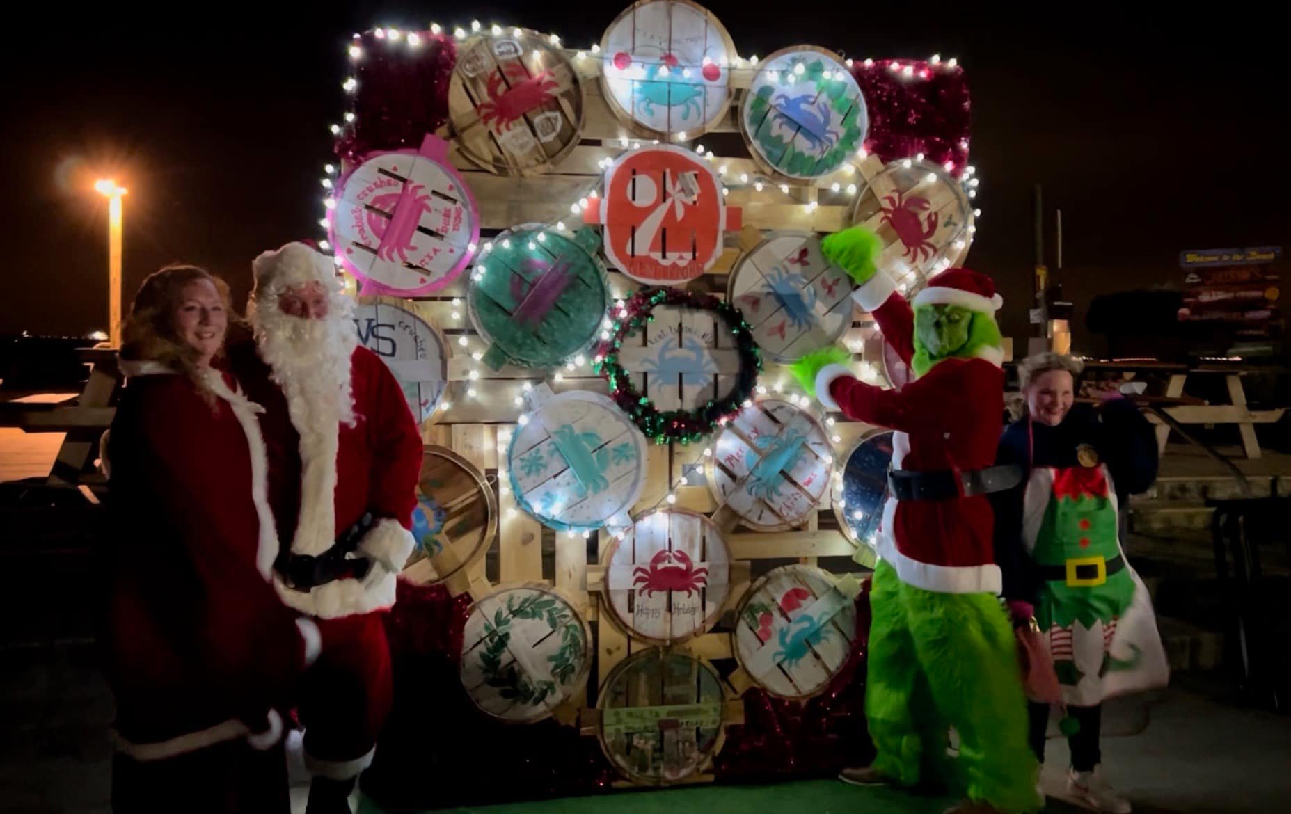 Four people in Christmas costumes, including Santa, Mrs. Claus, the Grinch, and an elf, pose in front of a festive, illuminated holiday display made of decorated barrels at night.