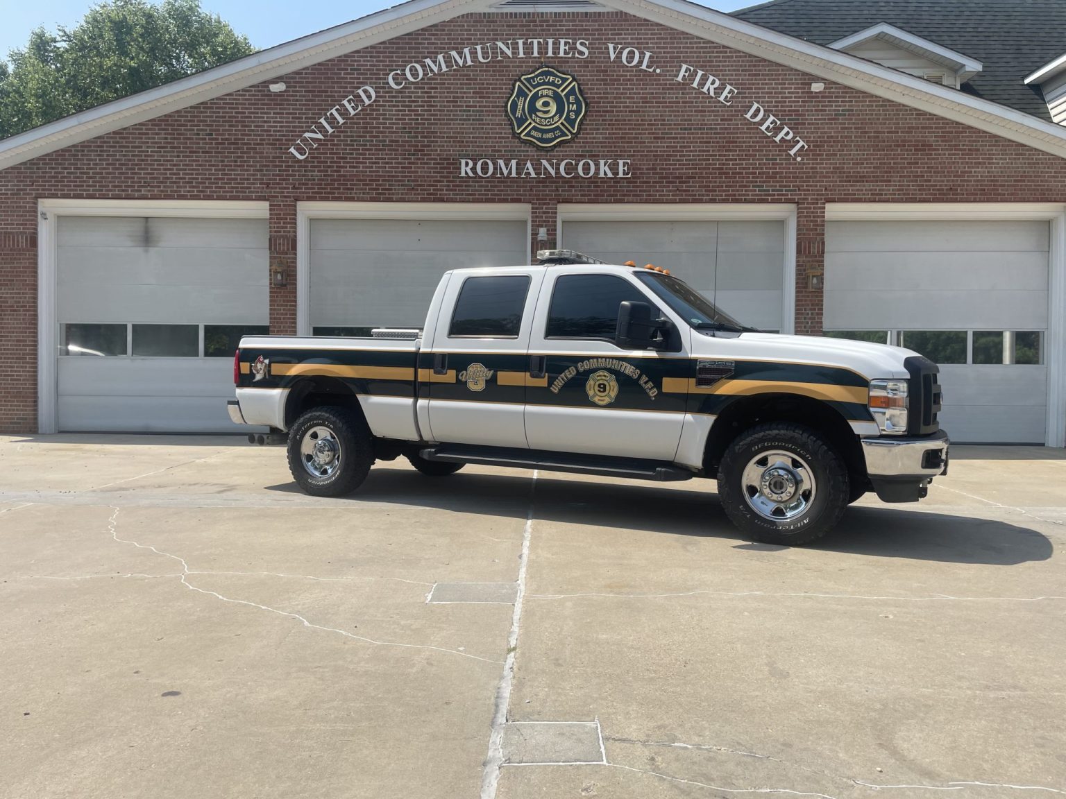 A white pickup truck with green and gold decals labeled "United Communities Volunteer Fire Dept" is parked in front of a brick fire station with three garage doors and signage reading "RomanCoke.