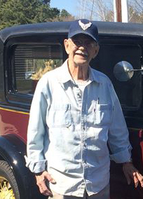 An elderly man wearing a blue cap and light denim shirt stands in front of a vintage car on a sunny day, smiling at the camera.