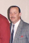A man with short hair and a mustache is wearing a gray plaid suit, a white shirt, and a tie. He is standing indoors against a plain light-colored wall and looking at the camera.