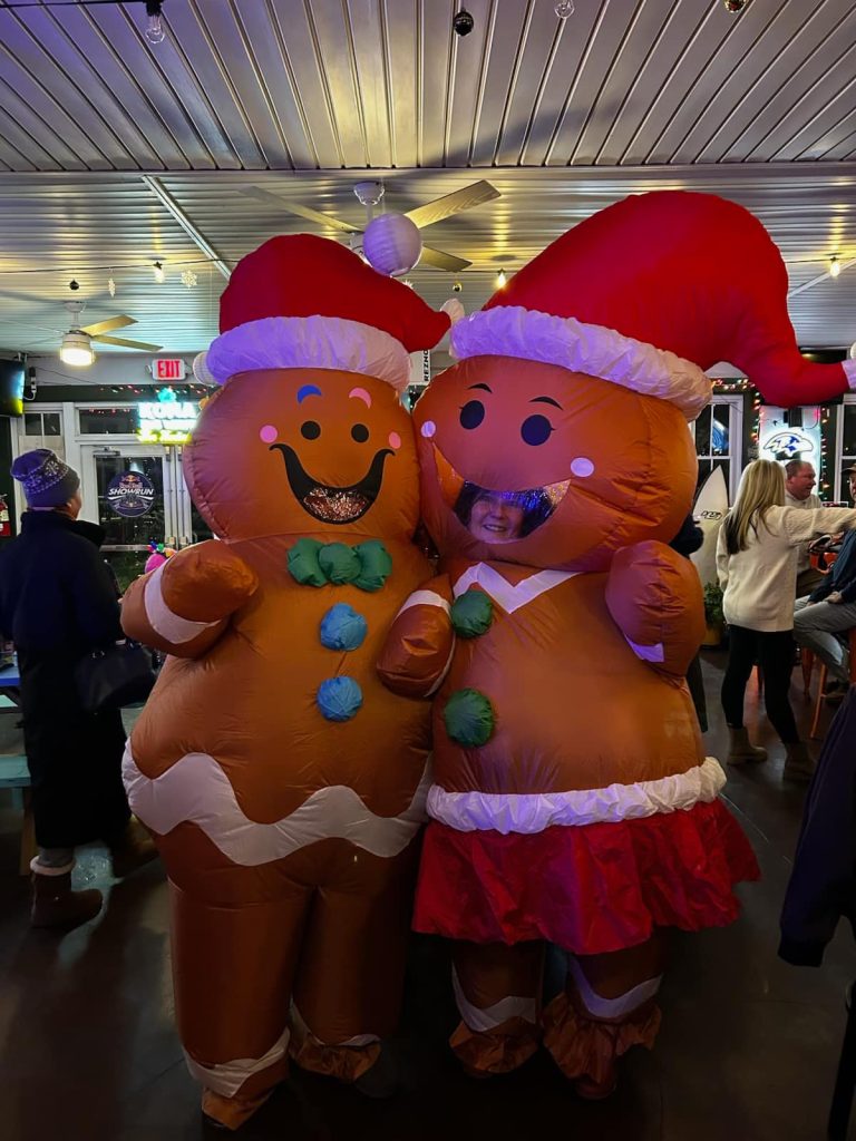 Two people dressed in large inflatable gingerbread costumes with Santa hats stand indoors at a festive event, smiling. Other people and holiday decorations are visible in the background.