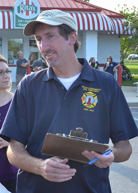 A man in a navy blue polo shirt and beige cap holds a clipboard and pen outside Rita’s Ice. People stand and sit in the background under a red-and-white striped awning.