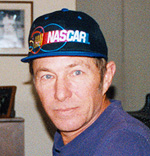 A man wearing a blue NASCAR cap and a purple shirt looks at the camera indoors, with framed photos and furniture visible in the background.