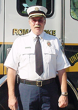 A uniformed fire chief stands in front of a fire truck with part of the word "Roanoke" visible on the vehicle behind him. He is wearing a white shirt, black tie, and a white peaked cap.