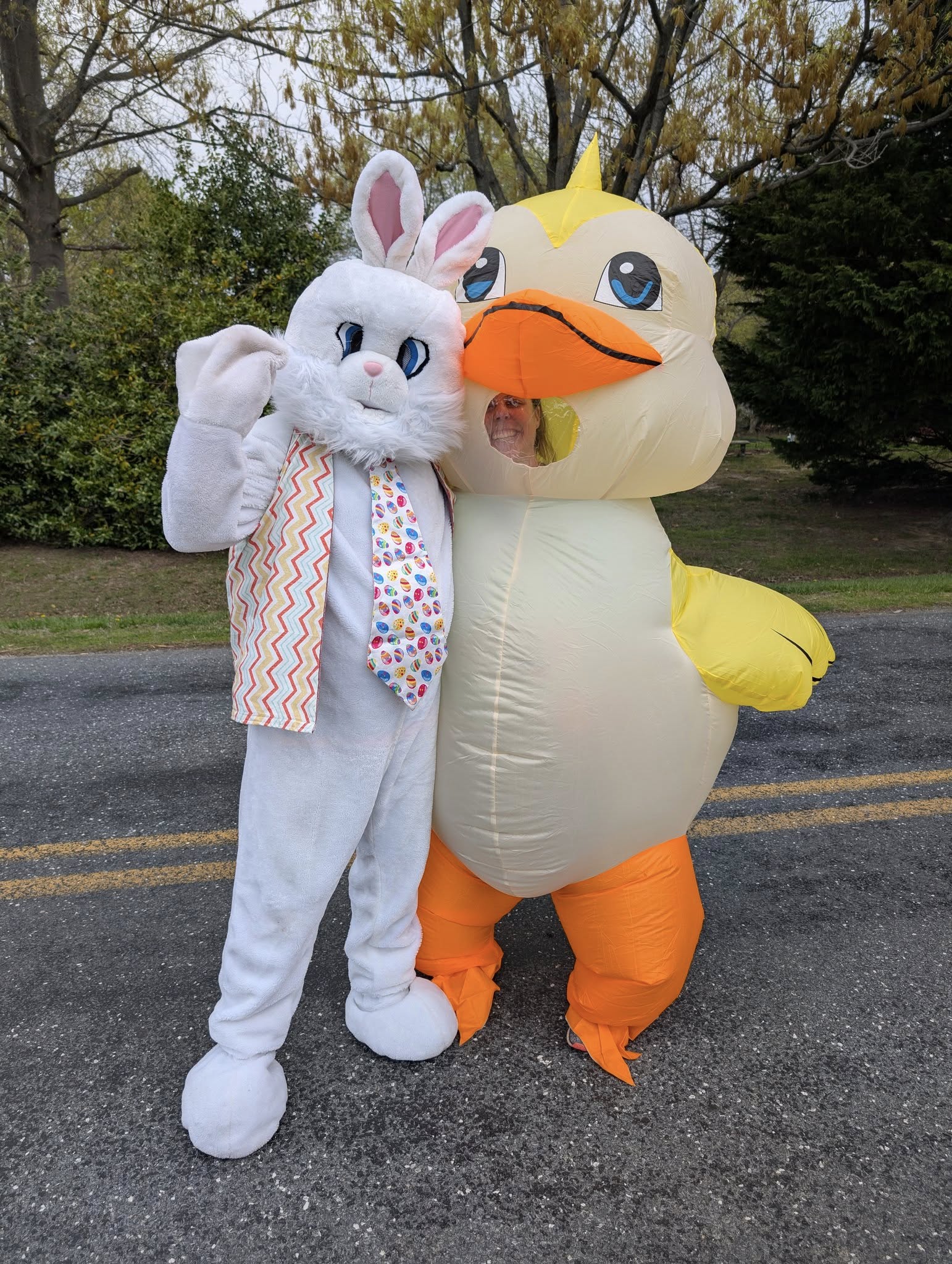 Two people in costumes stand on a road: one is dressed as a white Easter Bunny with a colorful tie, and the other is in an inflatable yellow duck costume, both posing cheerfully outdoors.