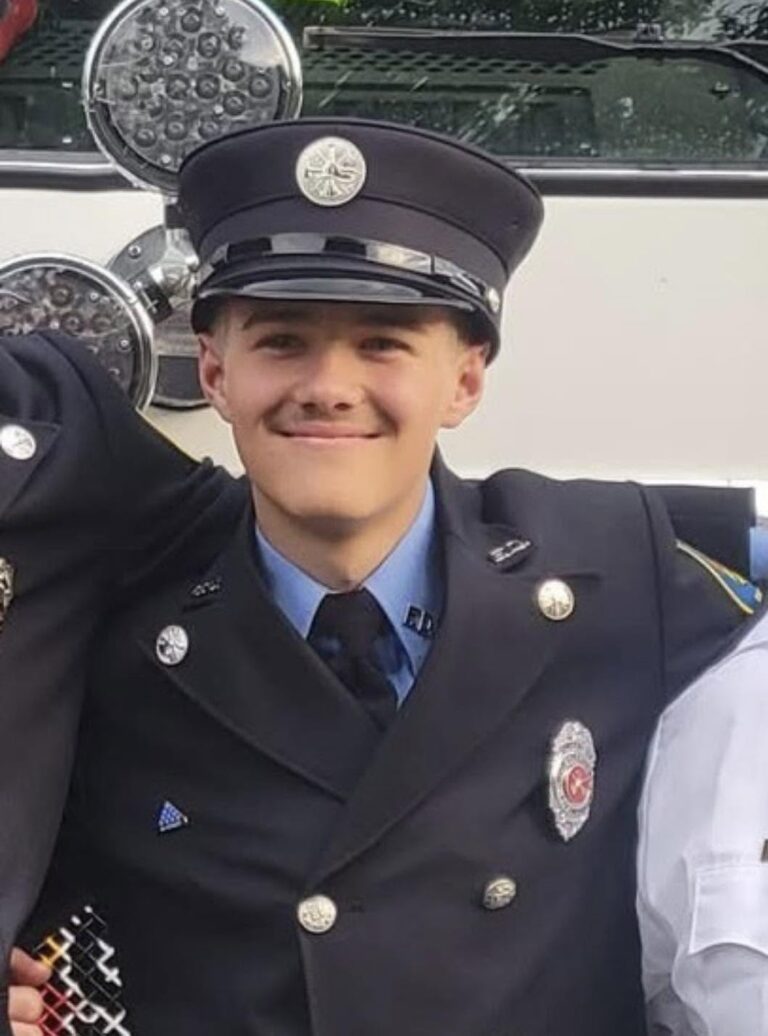 A young firefighter in uniform and cap smiles at the camera, standing in front of a fire truck with arms around others who are partially visible at the edges.
